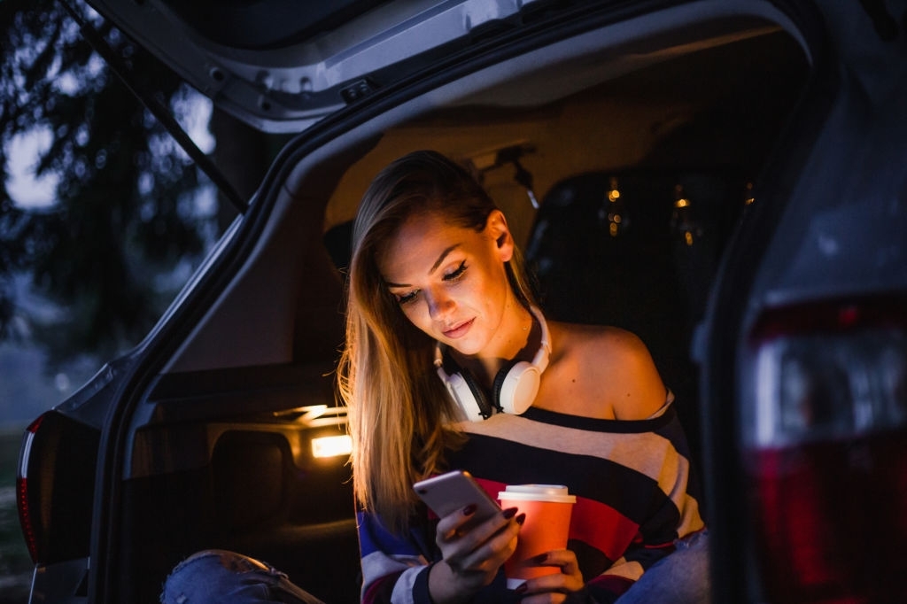 A young woman relaxes camping, sits in the back of a car surrounded by decorative light bulbs and enjoys the beauty of a warm spring night.
