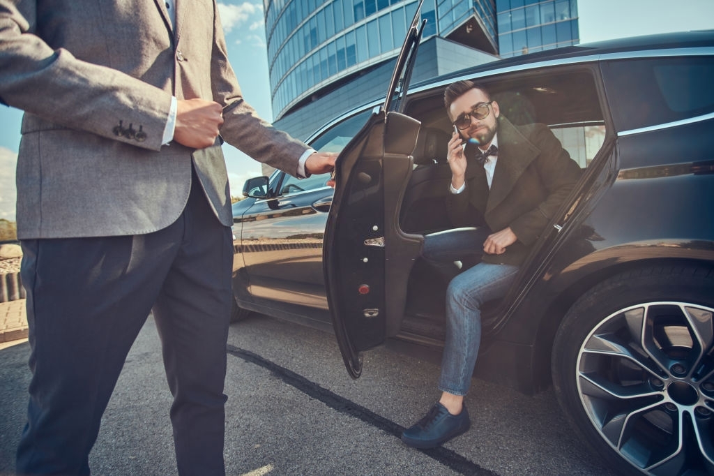 Attractive groomed man in sunglasses is talking by smartphone and sitting in the car while his assistant is opening door for him.